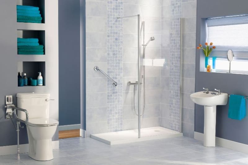 Accessible bathroom with grab bars, shower, pedestal sink, and toilet. Teal accents on a gray and white tiled backdrop.