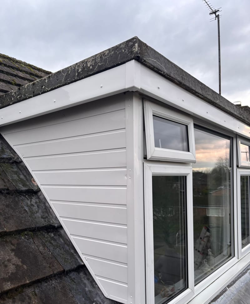 A white dormer window with horizontal cladding and white trim, viewed from the side against a roof with dark tiles.