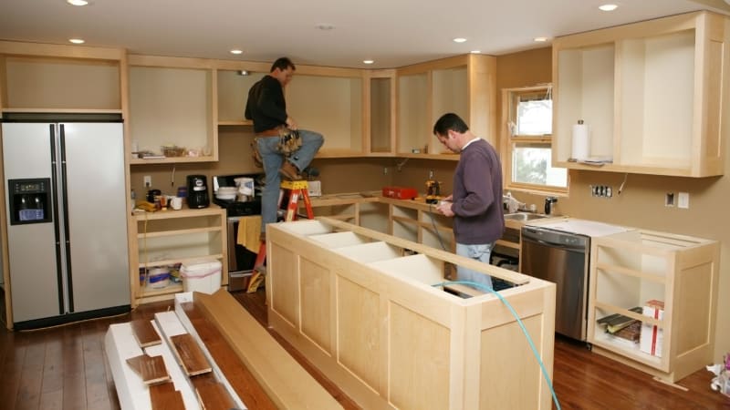 Two men installing kitchen cabinets in a kitchen with new wood floors and a stainless steel refrigerator.