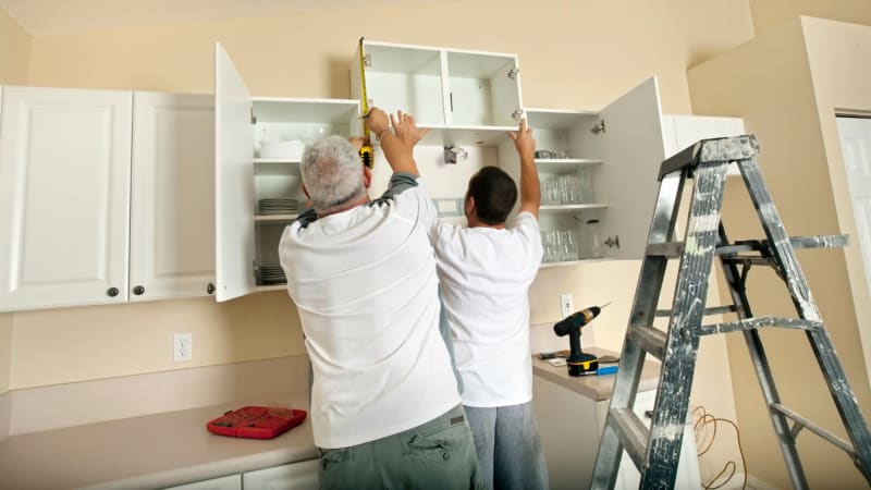 Two men installing white kitchen cabinets with a tape measure and drill.