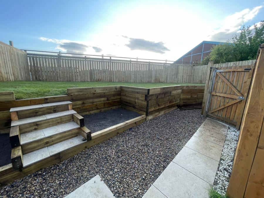 A multi-level backyard with wooden retaining walls, a staircase leading to a grass area, and gravel ground cover.