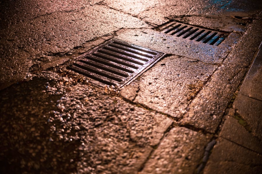 Rain-slick sidewalk with two metal storm drains at night