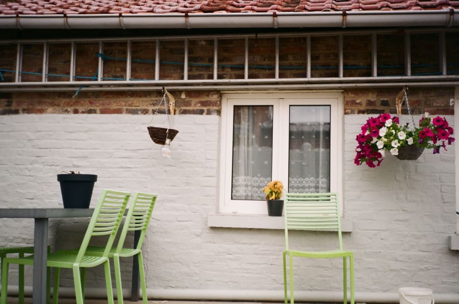 White patio with lime-green chairs, table, window, and hanging pink flowers on a brick wall.