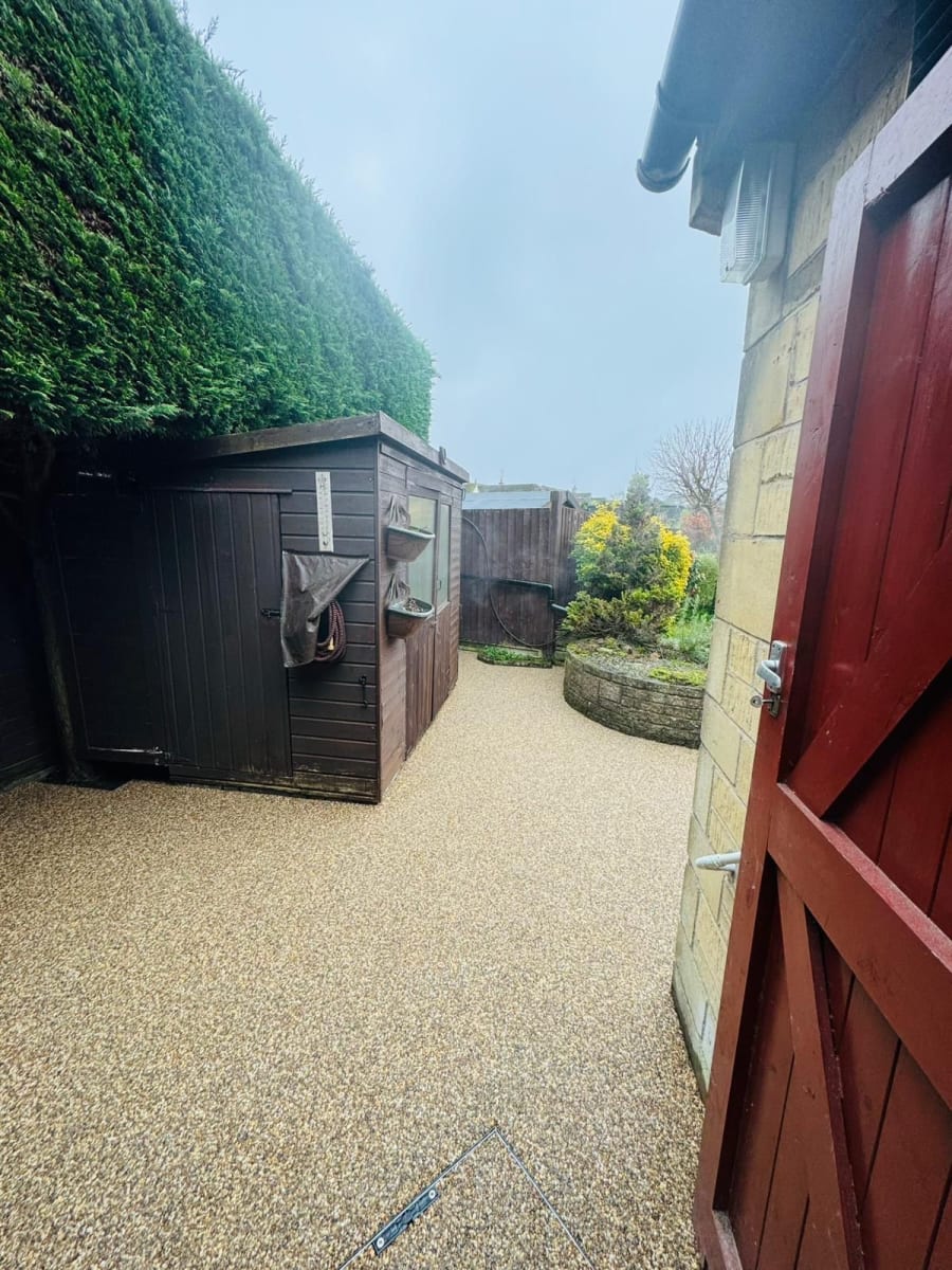 A gravel path leads between a dark wooden shed and a stone wall, viewed through an open red gate.