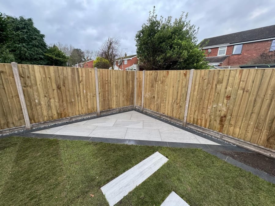 Backyard corner with wooden fence, concrete pad, and a white plank on the grass.