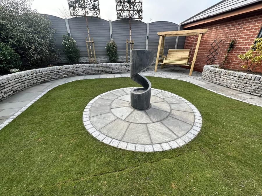 Circular stone patio with a black spiral sculpture, surrounded by a curved lawn and brick garden wall