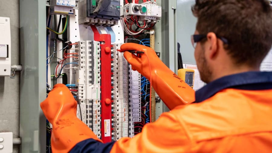 Technician in orange gloves inspecting electrical panel wiring in a control cabinet