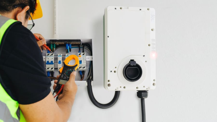 An electrician in a high-visibility vest uses a clamp meter to test electrical components next to a wall-mounted EV charger.