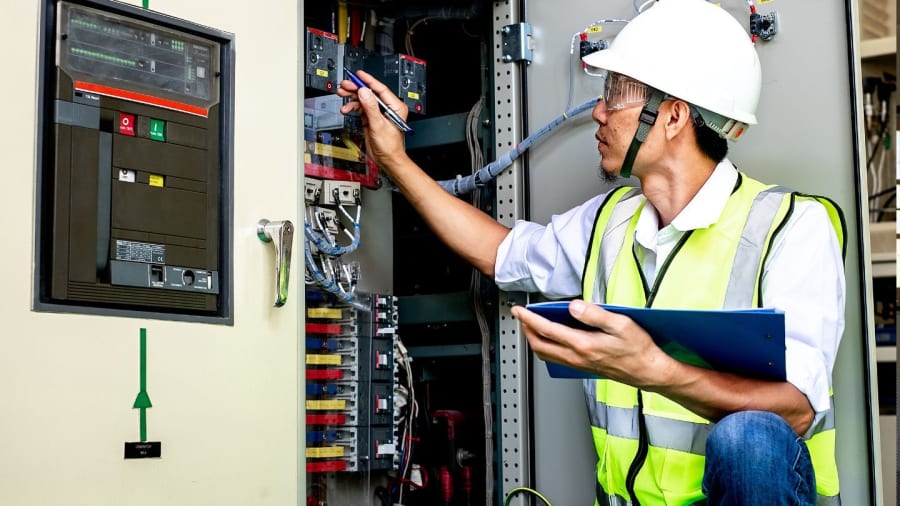 Technician in hard hat inspecting an electrical control panel with a tablet in a utility room