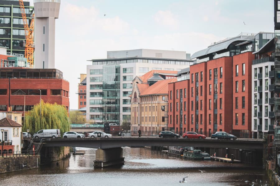 Urban riverfront with a bridge, modern buildings, and a red brick waterfront under a cloudy sky
