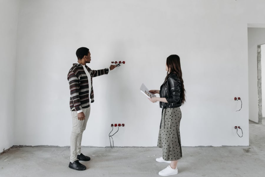 Two people discussing wall outlets in a bare white room, one pointing at wiring.
