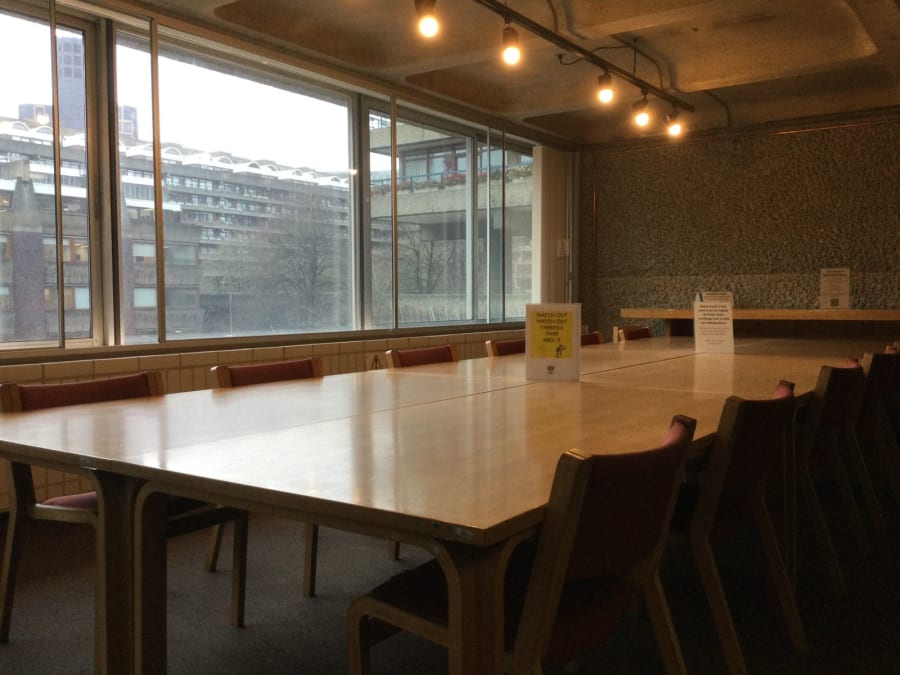 Empty conference room with long wooden table, chairs, and large windows overlooking buildings