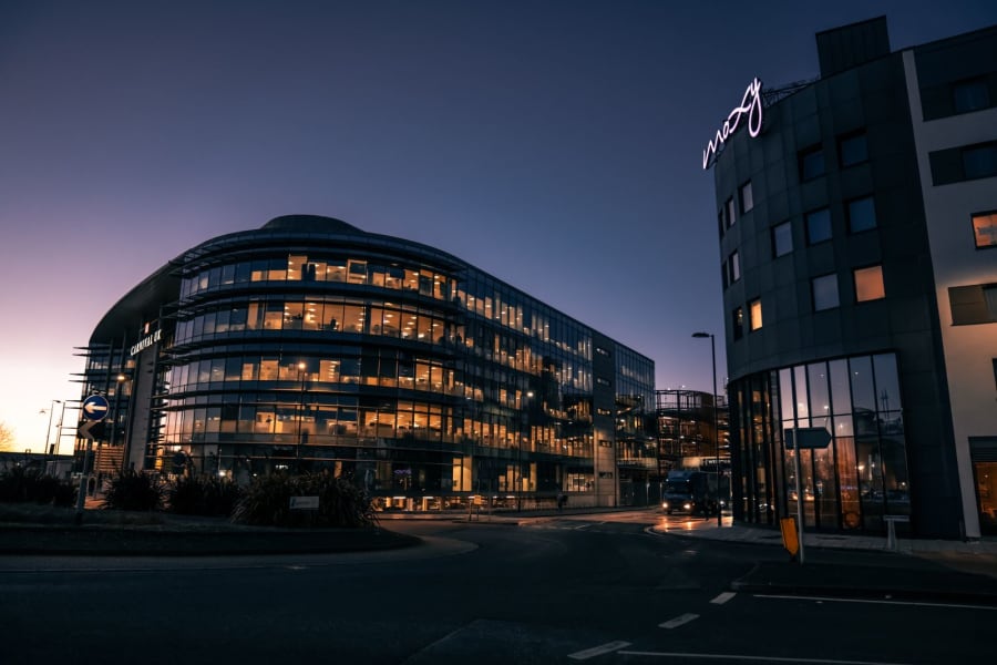 Dusk view of a modern glass office building lit from within, with a city street in the foreground