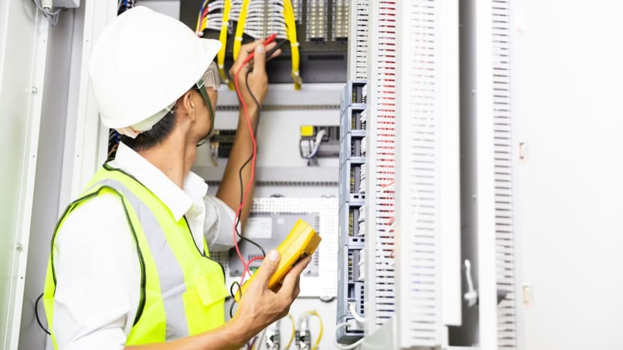 A technician in a white hard hat and yellow safety vest uses a multimeter to test electrical wires in a control panel.