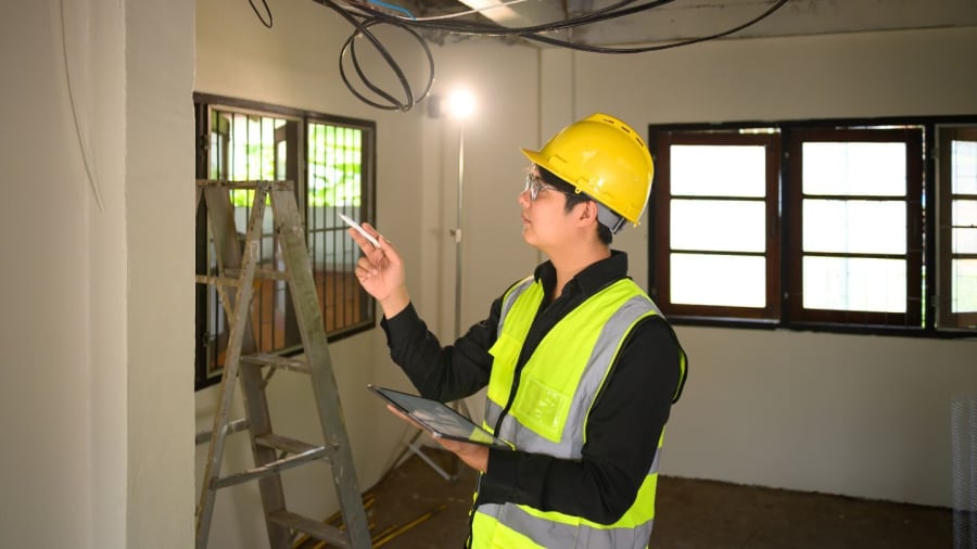 A technician in a white hard hat and yellow safety vest uses a multimeter to test electrical wires in a control panel.