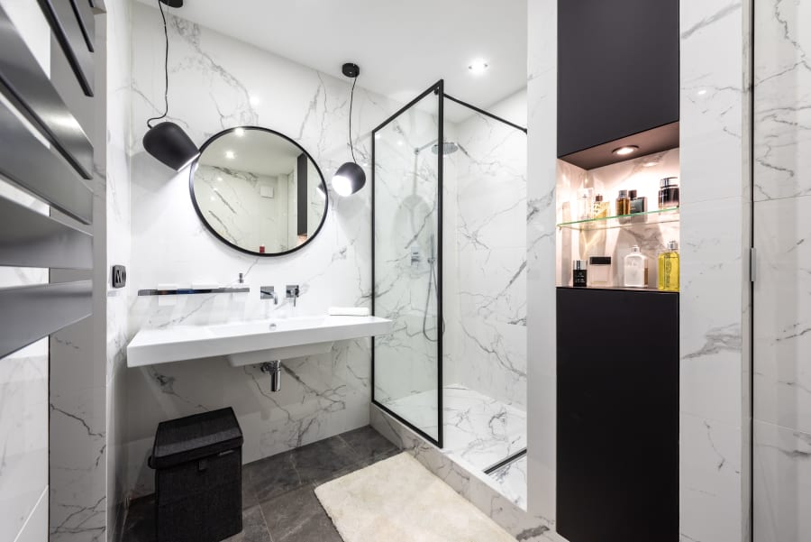 Modern bathroom with marble walls, round mirror, white vanity, glass shower, and black trash bin
