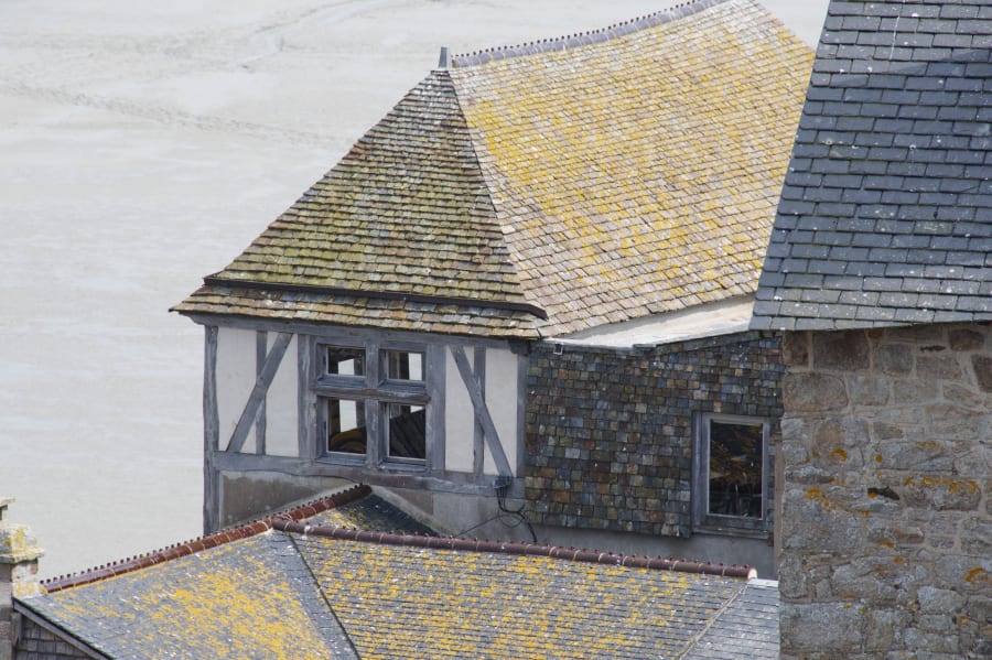 Stone and timber house with gray and yellow shingled roofs, seen from above against a pale sky