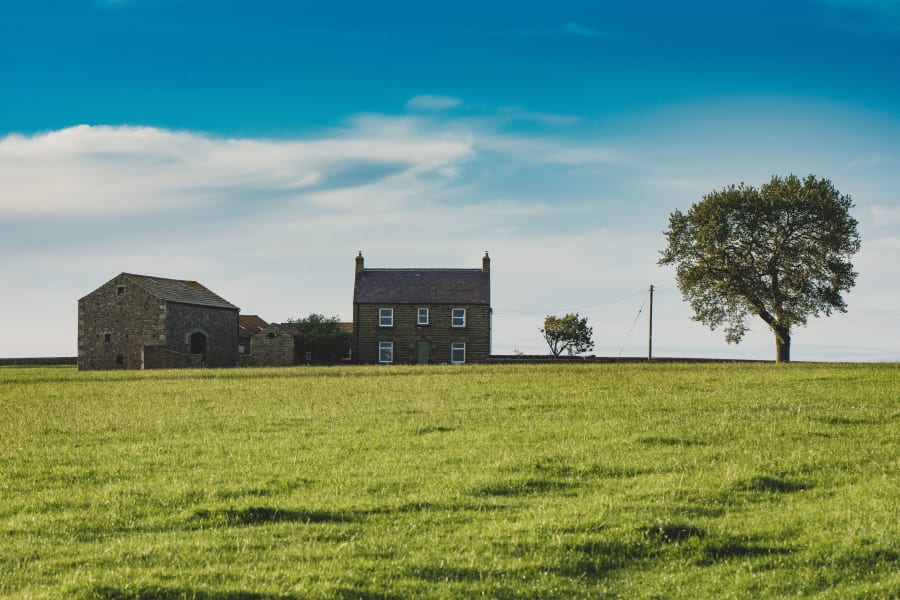 Rural farmhouse and barn beside a lone tree in a wide green field under a blue sky