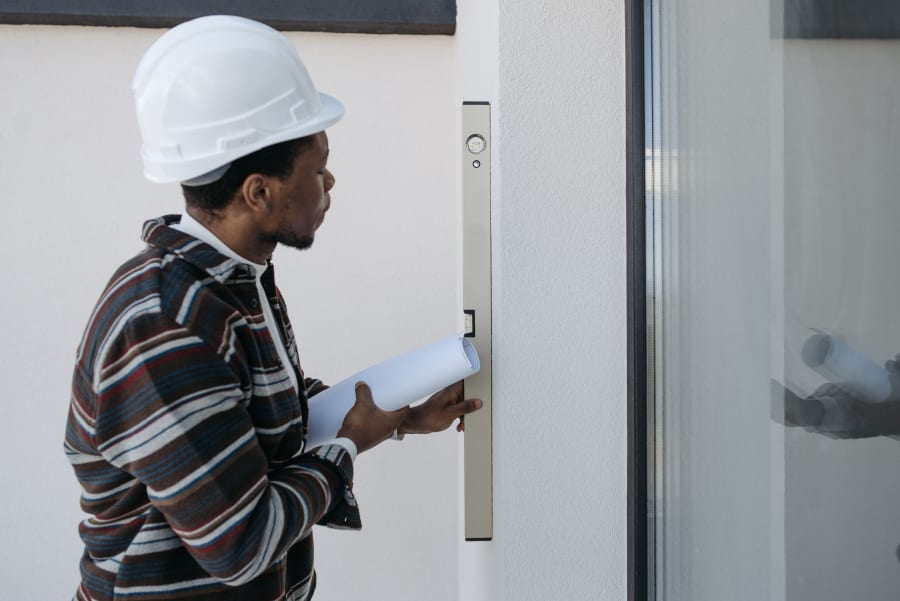 Worker in a white hard hat holding papers beside a glass door and wall-mounted access panel