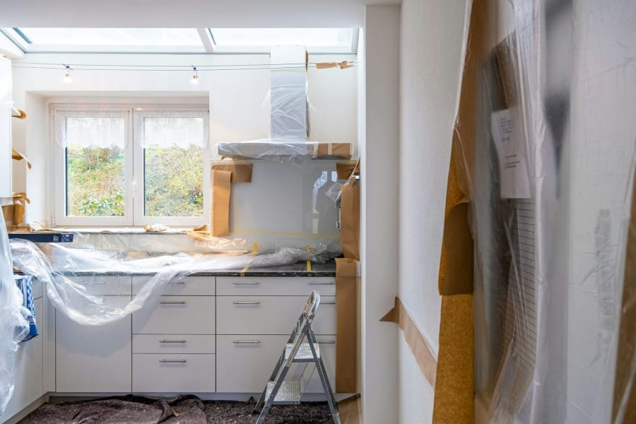 Kitchen under renovation, white cabinets, plastic sheeting, ladder, window, exposed walls.