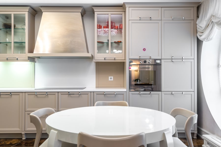Bright modern kitchen with gray cabinets, white counters, built-in oven, and round white table by the window.