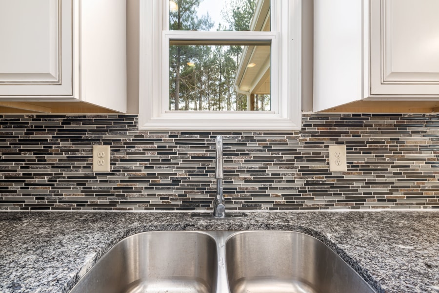 Kitchen sink beneath window with gray granite countertop and mosaic tile backsplash