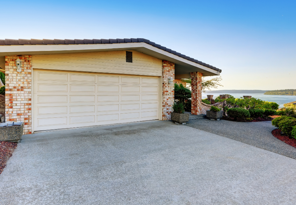 Garage with concrete driveway, beige garage door, and stone accents under a blue sky near water.