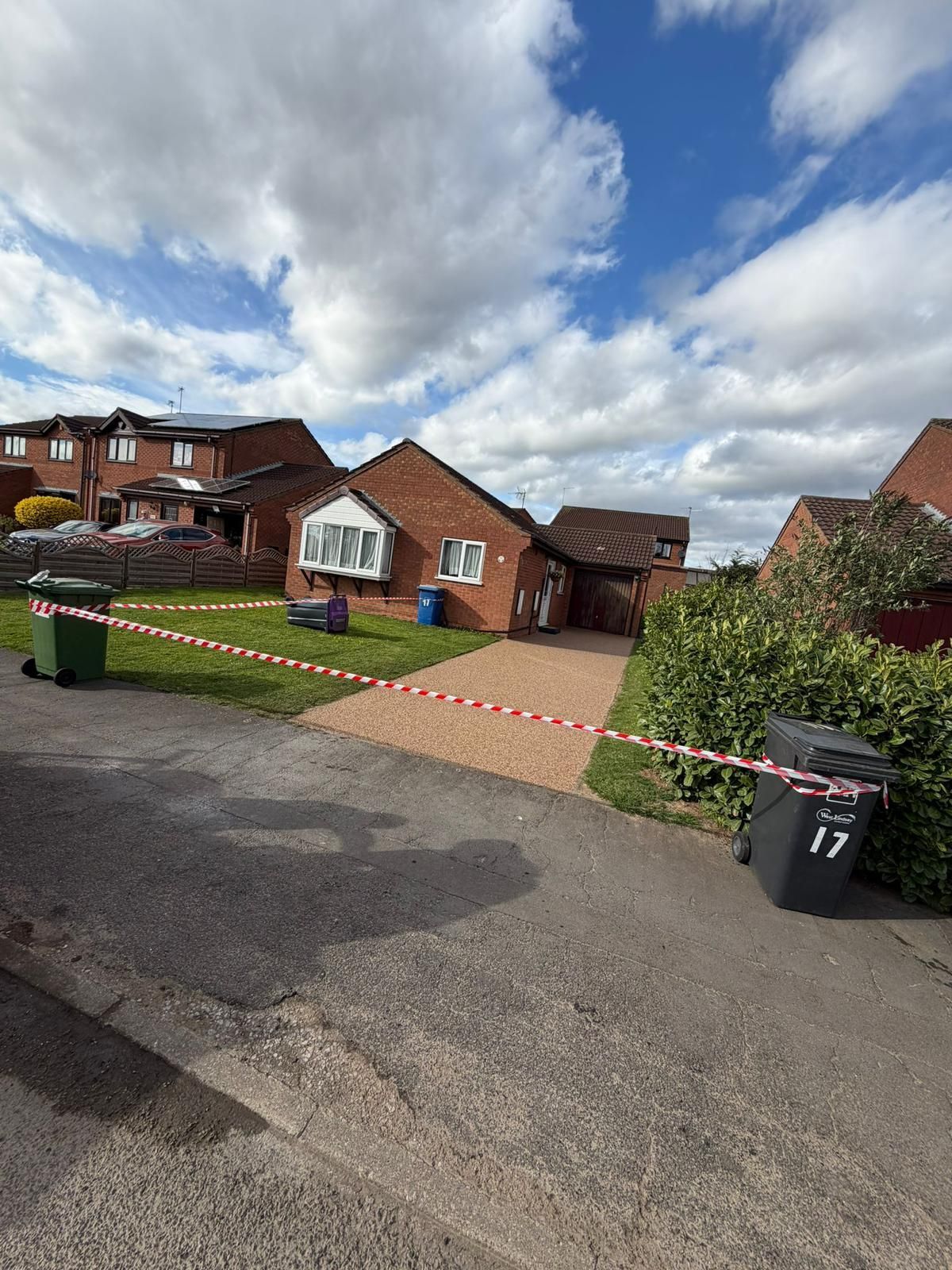 A single-story brick house with a paved driveway cordoned off by red and white hazard tape on a sunny day.