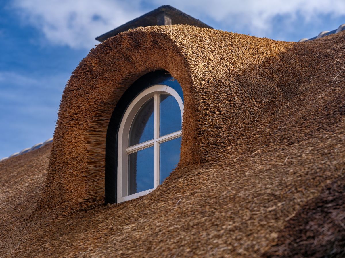 Round attic window set in a steep thatched roof under a blue sky