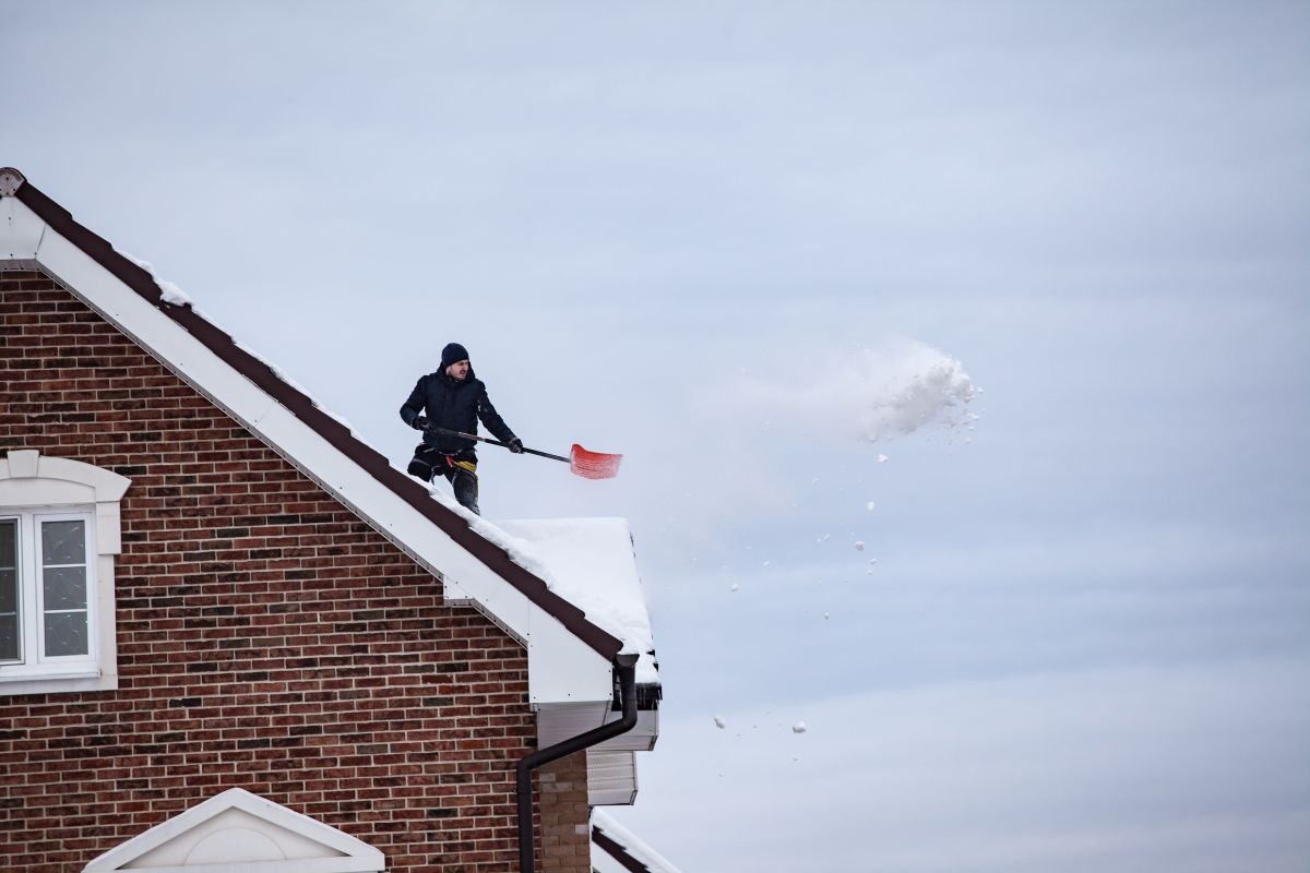 Person shoveling snow from a rooftop beside a brick house, with snow falling nearby