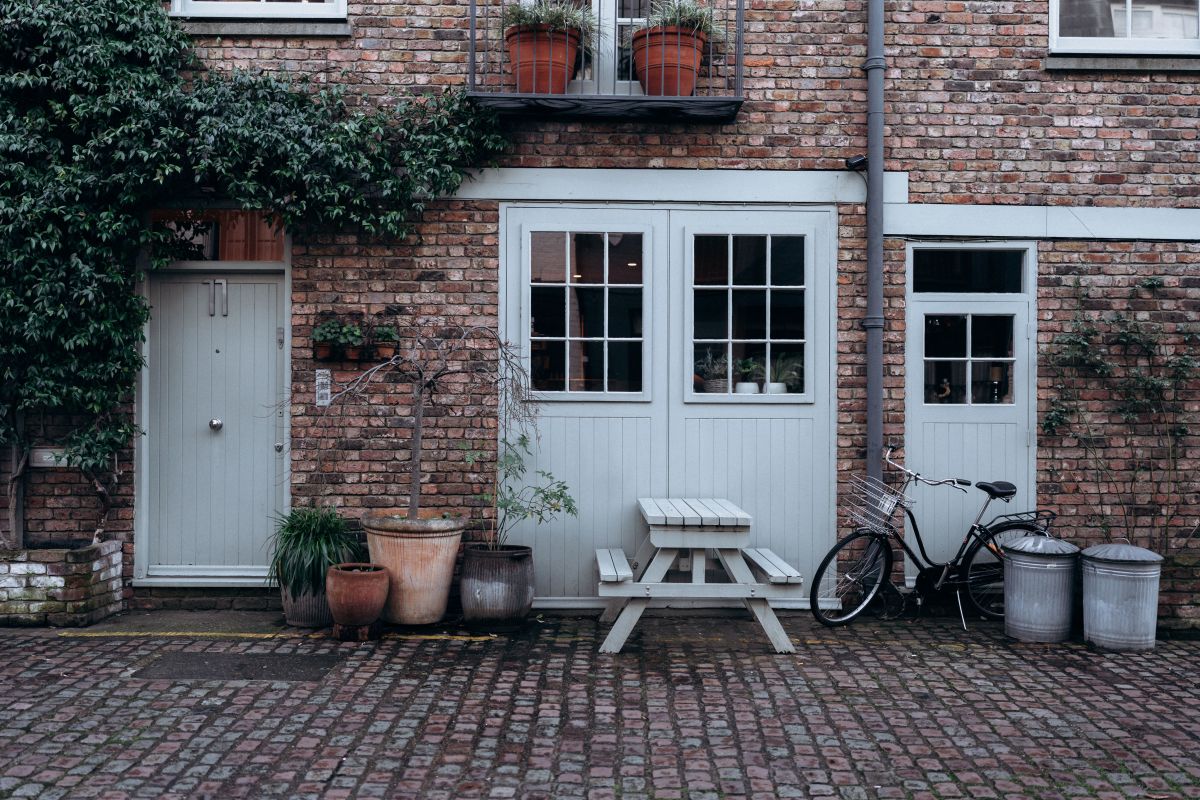 Brick townhouse facade with white doors and windows, potted plants, a bench, and a bicycle on cobblestones