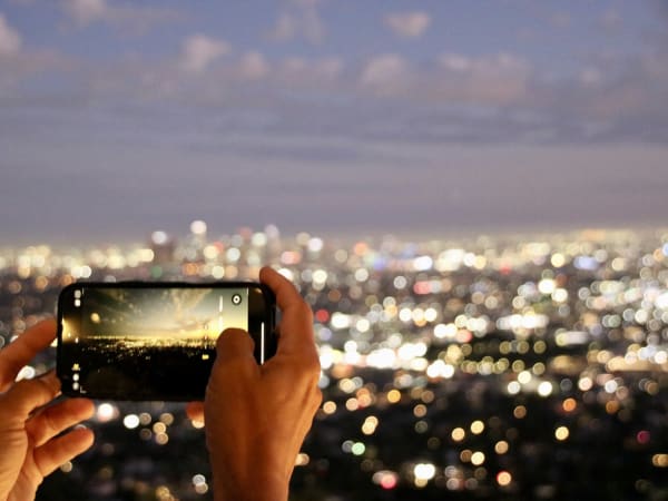 Hands holding a smartphone to photograph blurred city lights at Griffith Observatory, USA.