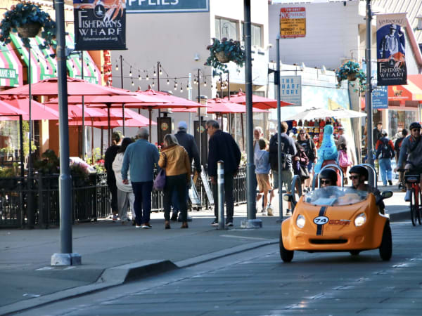 San Francisco waterfront street with orange rental vehicle, red umbrellas, and pedestrians