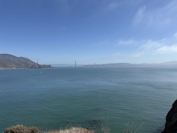 Wide bay view in San Francisco with distant bridge, hillsides, and rocky coastal vegetation
