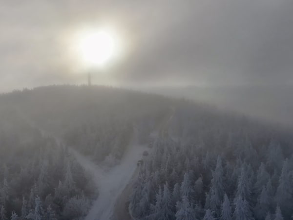 Snow-covered forested hillside with fog and tower silhouette at Fichtelberg Sa.