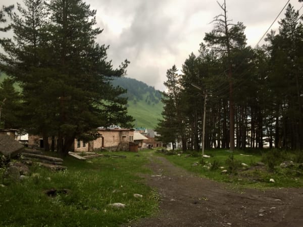 Dirt track through mountain settlement with stone buildings and conifers, Mount Elbrus Expedition