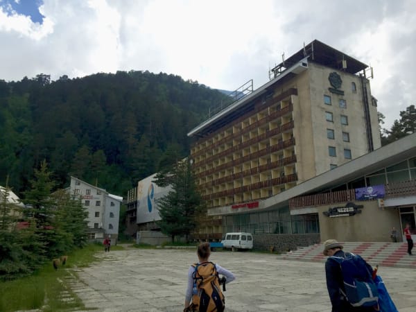 Two hikers with backpacks crossing concrete plaza toward Soviet-era hotel, Mount Elbrus Expedition