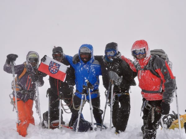 Five mountaineers in winter gear holding a flag at snowy Mount Elbrus Expedition summit