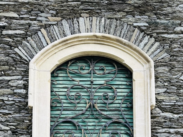 Arched stone window with wrought iron grille and green shutters in Verbania