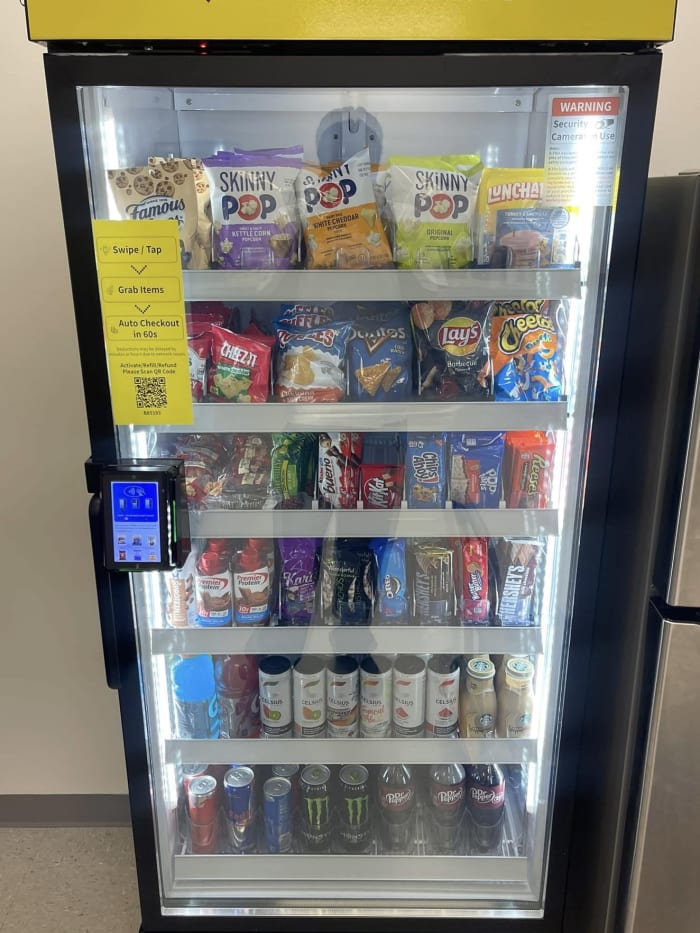 Yellow-header managed micro market unit stocked snack-forward with SkinnyPop kettle corn, Cheez-It, Doritos, Lays, Cheetos, Snickers, Reese's, and Hershey's bars.