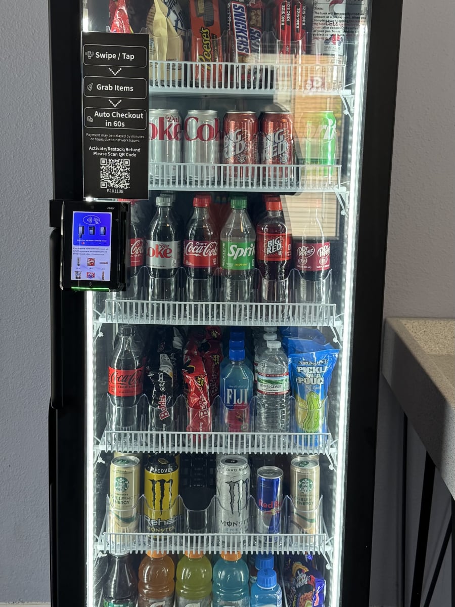A floor-to-ceiling managed micro market unit installed in a workplace, fully stocked with sodas, energy drinks, water, snacks, and grab-and-go essentials. Branded '24H Smart Vending' with a built-in tap-to-pay screen on the side.