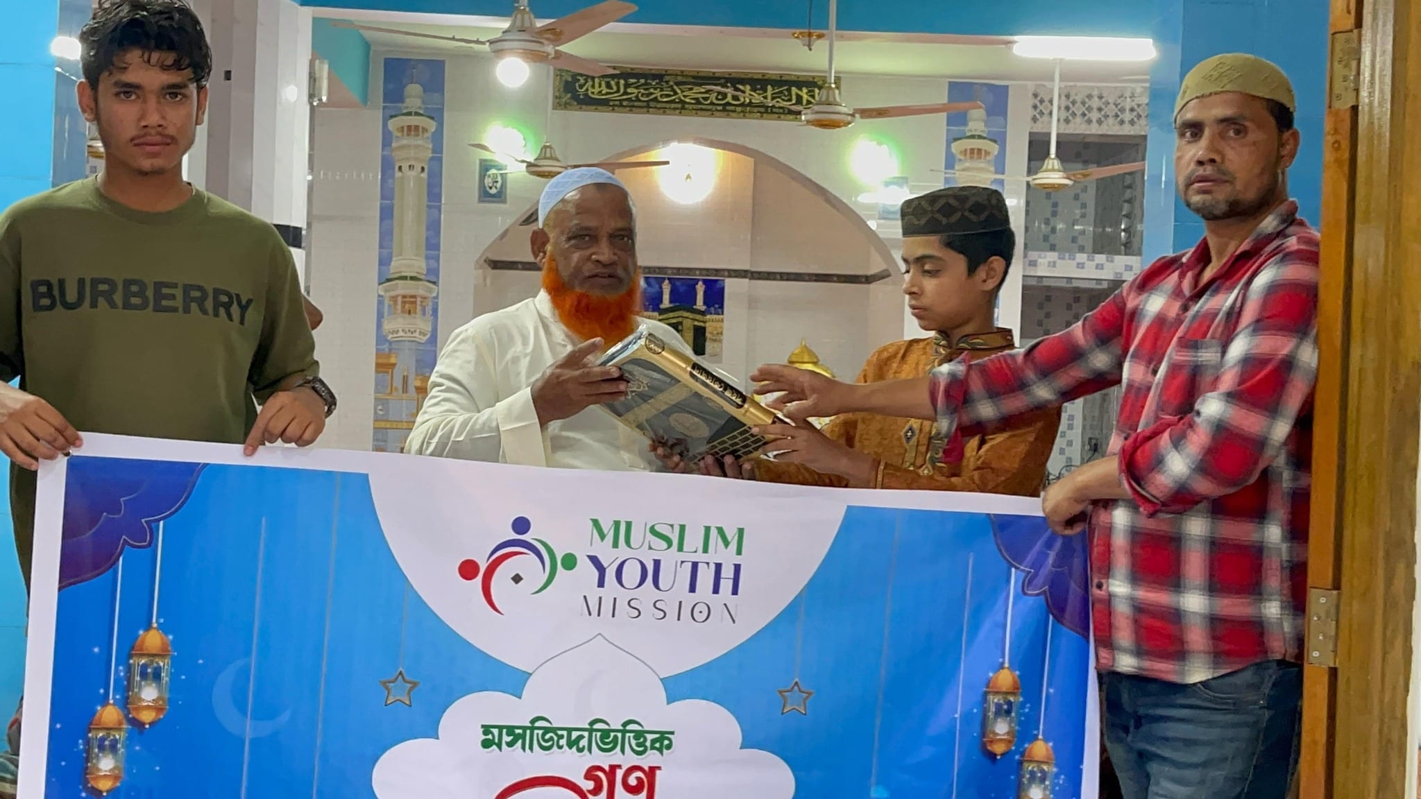 Four men stand beside a blue banner with a mosque design, likely a Muslim Youth Mission promotional image.