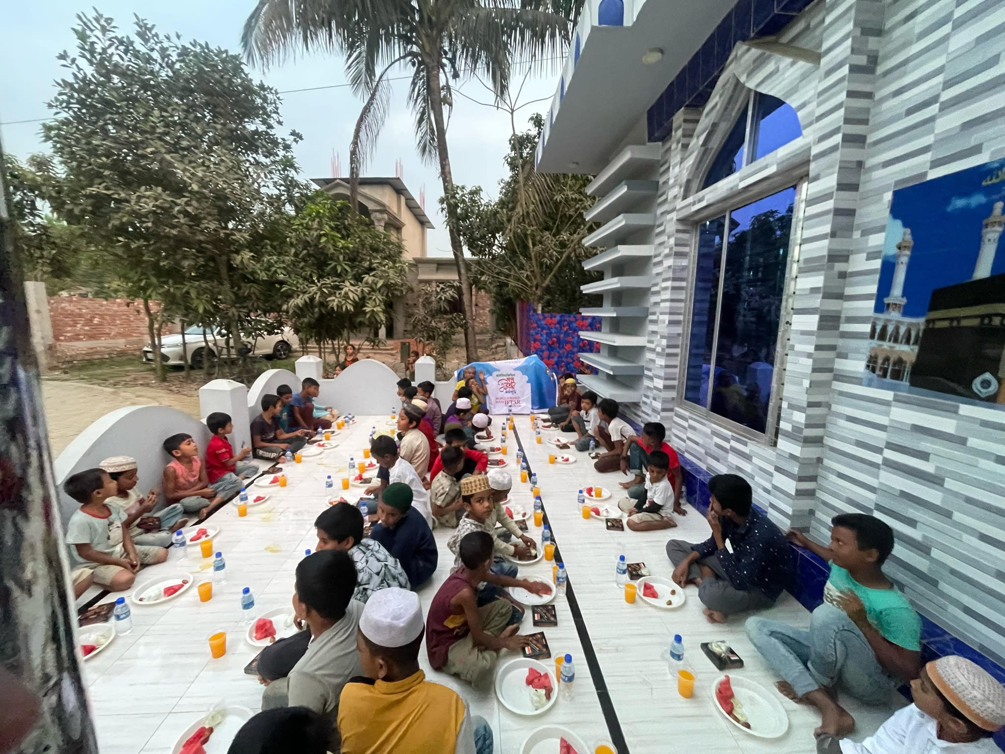 A large group of people, mostly children, are seated at a long table enjoying a meal together in an outdoor setting with palm trees and buildings in the background.