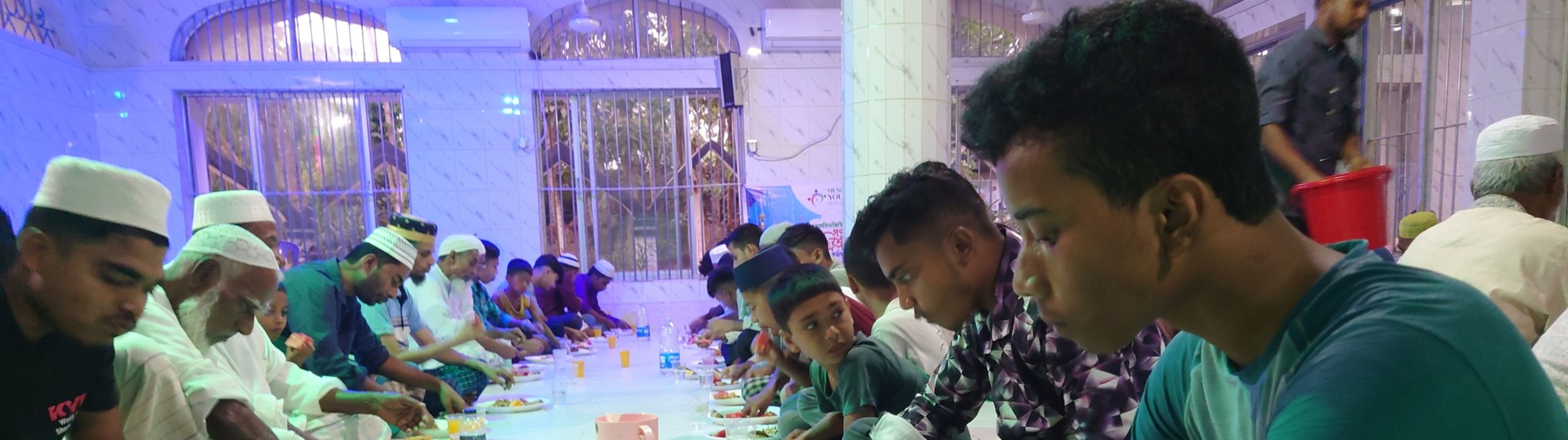 Several men are seated on the floor, eating and drinking, in a brightly lit room.