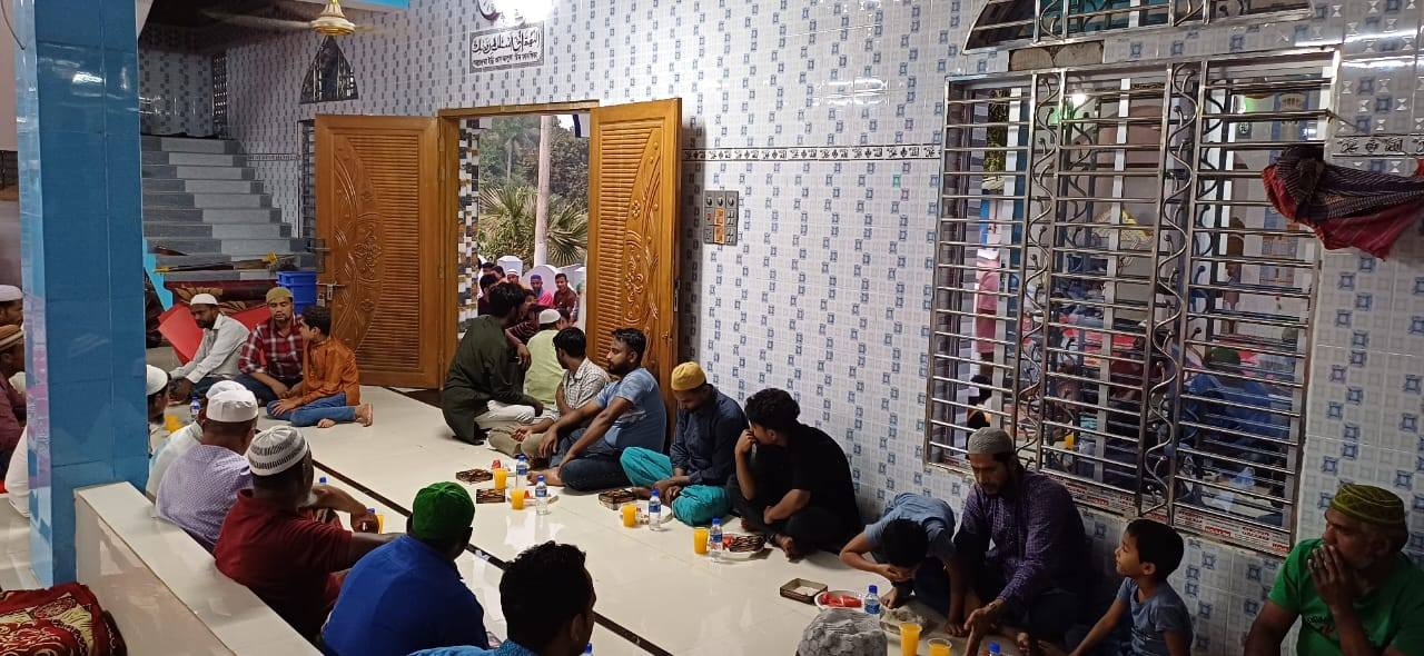 A crowded indoor space with people sitting on the floor, surrounded by colorful walls and windows, suggesting a community gathering or religious setting.