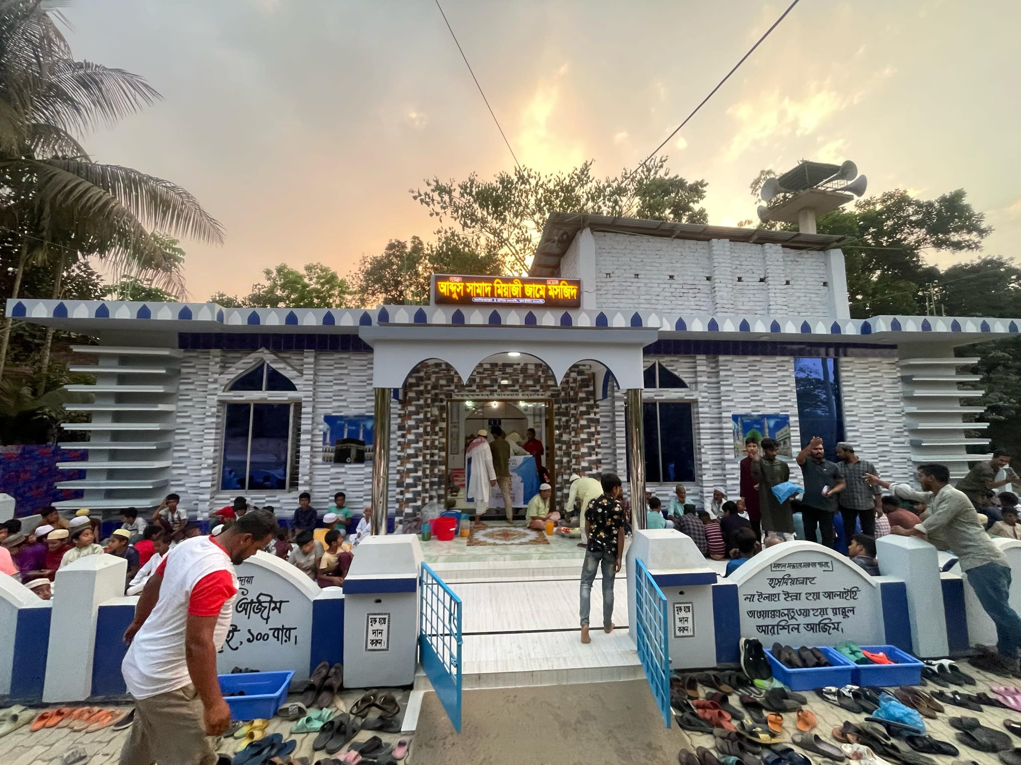 A colorful and ornate mosque structure surrounded by a crowd of people, set against a backdrop of trees and a bright sky.