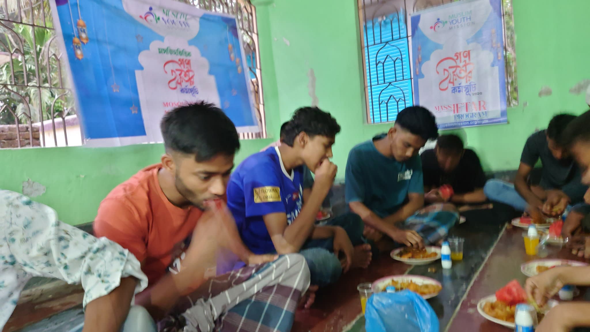 The image shows a group of people, likely students, sitting on the floor and eating a meal together in what appears to be a classroom or community space with colorful walls and posters in the background.