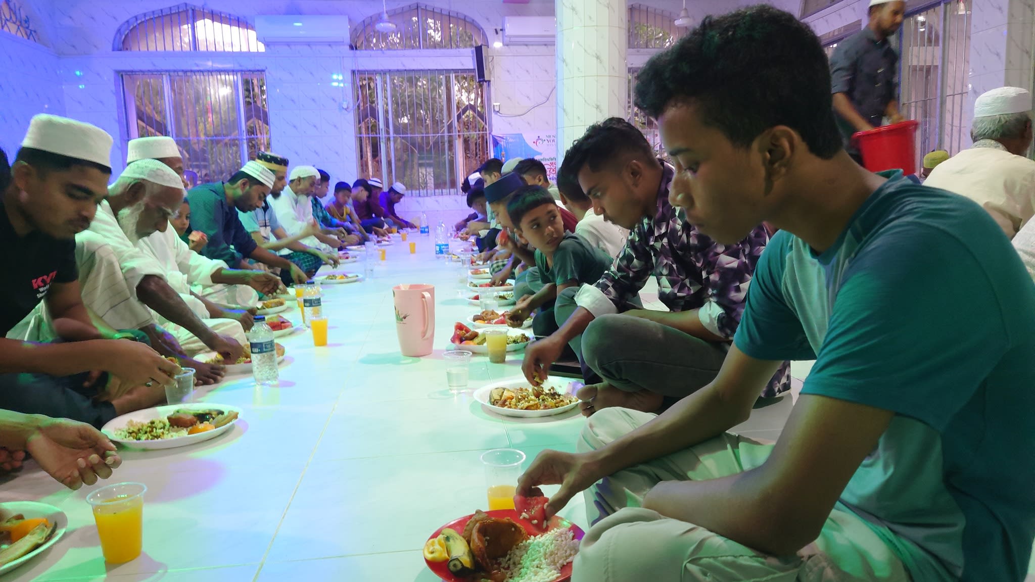 Several men are seated on the floor, eating and drinking, in a brightly lit room.