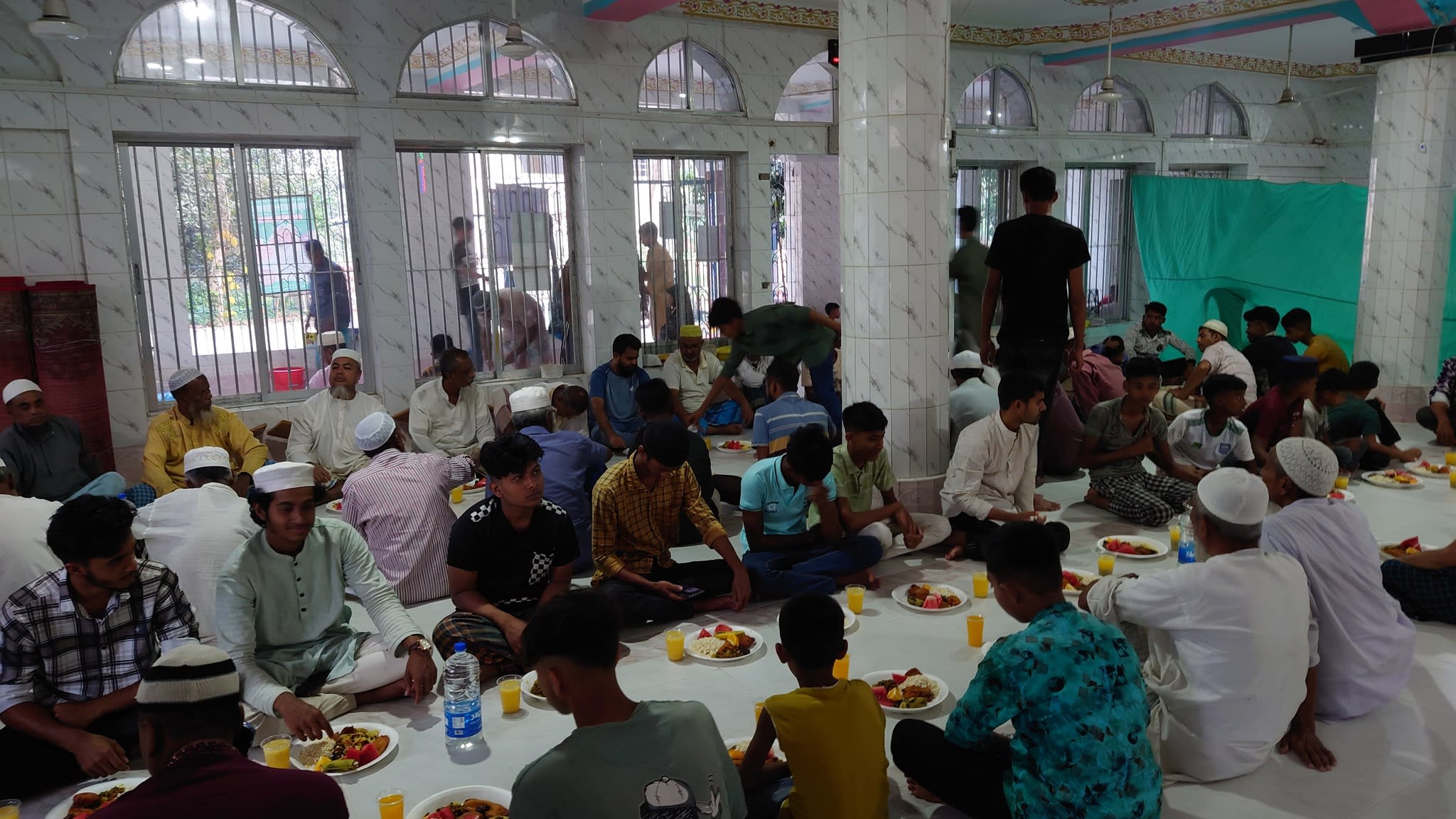 Many men seated on the floor in a hall, likely for a meal or gathering.