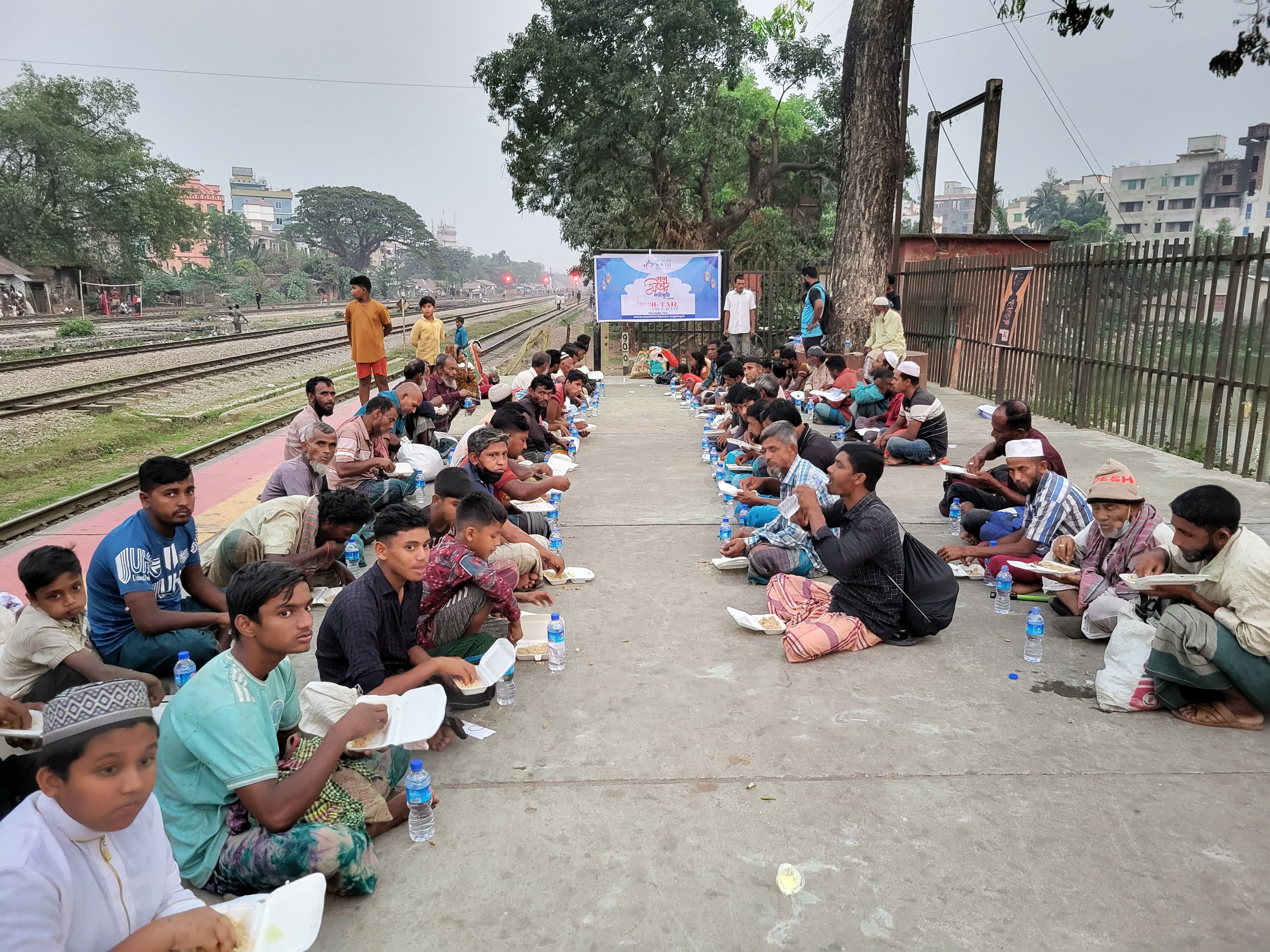 A large group of people, mostly men, sitting on the ground along a railway track, surrounded by trees and buildings in the background.